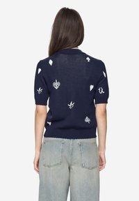 Námořnicky modrý krátký svetr s bílými vyšívanými tvary, včetně srdcí a listů, s vroubkovaným lemem.