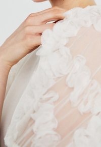 Detailní záběr ruky, která se dotýká límce transparentního bílého volánkového oděvu v oblasti horního hrudníku a krku osoby.