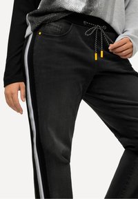 Pantalones de chándal negros estilo jogger hechos de denim, con una cintura elástica negra y cordones ajustables. Rayas laterales en blanco y detalles en amarillo.