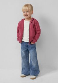 Rote Kapuzenjacke mit Frontreißverschluss, weißes T-Shirt und ausgestellte blaue Jeans mit floralen Stickereien, kombiniert mit hellen Sneakers.