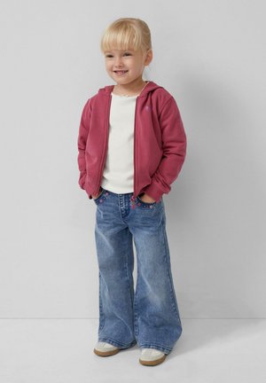 Rode hoodie met een rits aan de voorkant, wit shirt en uitlopende blauwe jeans met bloemborduursels, gecombineerd met lichtgekleurde sneakers.