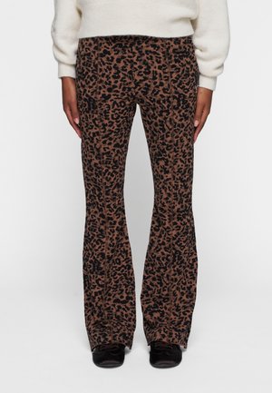 Persoon in bruin en zwart luipaardprint flared broek, een witte trui en zwarte schoenen, staande tegen een effen witte achtergrond.