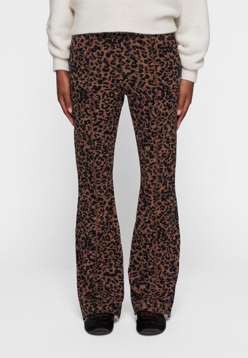 Person iført brune og sorte leopardprint flared bukser, en hvid sweater og sorte sko, der står foran en ensfarvet hvid baggrund.