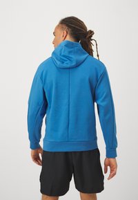 Hoodie azul de algodão com uma textura suave, apresentando um capuz grande, punhos e bainha canelados, e um design minimalista, usado sobre calções pretos.