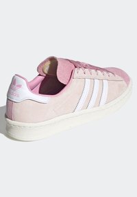 Růžové semišové tenisky s bílými koženými pruhy, polstrovaným jazykem a bílou gumovou podrážkou. Obsahuje vyražené logo na patě.