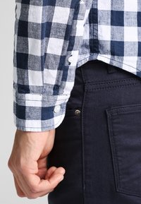 Kostkovaná modro-bílá košile s knoflíkovými manžetami, vystavená na tmavě námořnických kalhotách. Detailní záběr ruky na bocích.