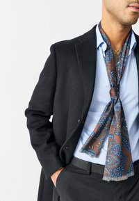 Schwarzer Wollmantel über einem hellblauen Hemd, mit einem gemusterten braun-blauen Schal, der am Hals gebunden ist. Schlanke schwarze Hose sichtbar.