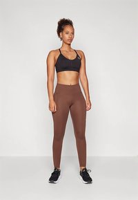 Črni atletski športni modrček in rjave pajkice z visokim pasom, z gladko teksturo in prileganim dizajnom. Model nosi črne superge.