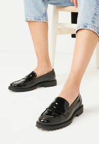 Zapatos de charol negro tipo slip-on con una suela gruesa, que presentan un acabado suave y una ligera textura. Punta redondeada y mínimos detalles decorativos.
