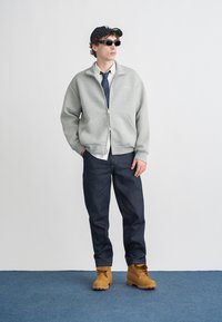 Sivá mikina na zips cez biele tričko, tmavomodré džínsy a žlté topánky. Doplnky zahŕňajú slnečné okuliare a čiernu pruhovanú čiapku.