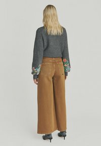 Brune brede bukser parret med en mørkegrå striktrøje med farverig blomsterbroderi på ærmerne.