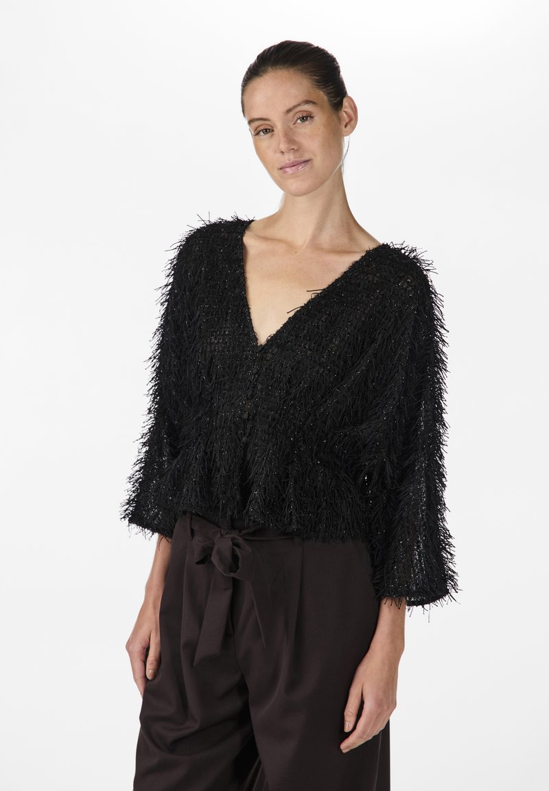 Blusa nera testurizzata a maniche corte con scollatura a V profonda e dettagli di frange. Indossata sopra pantaloni marrone scuro a vita alta con cintura a fiocco.