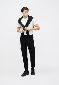 Sorte joggingbukser med cargolommer, kombineret med en lysegrå tætsiddende t-shirt. En sort sweater ligger over skuldrene. Sorte sneakers fuldender looket.