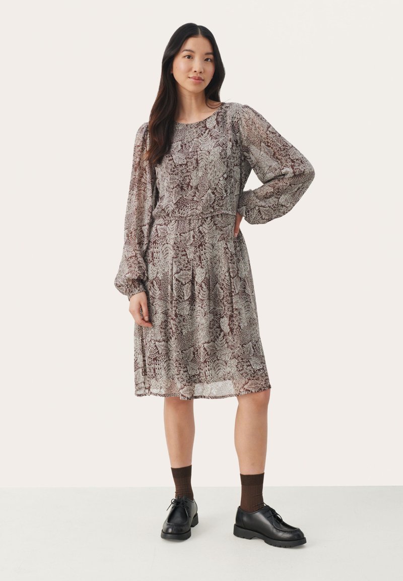 Langärmliges, knielanges Kleid in einem braun- und cremefarbenen Blättermuster, mit einer taillierten Taille und einem fließenden Rock. Leichtes, halbtransparentes Material.