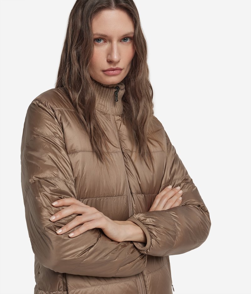 Frau mit langen braunen Haaren, die eine braune gesteppte Jacke mit Reißverschluss und einen gerippten Rollkragenpullover trägt, Arme verschränkt, neutraler Gesichtsausdruck.