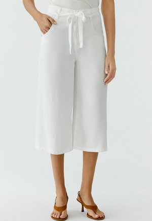 Frau trägt weiße, weit geschnittene, kurz geschnittene Hosen mit einem Stoffgürtel, der an der Taille gebunden ist, und braune Sandalen mit Absatz.