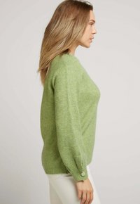 Pull en maille verte avec un col rond, de longues manches bouffantes et un accent de bouton au poignet. Fabriqué en tissu doux et texturé.