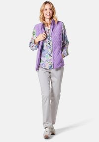 Lavendelfarbene, gestepptes Westenteil über einer mehrfarbigen, blumengemusterten Bluse, kombiniert mit hellgrauen Hosen und weißen Sneakers. Lässiges Outfit mit schichtweise gestaltetem Design.