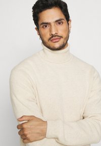 Beige coltrui gemaakt van een zachte, gestructureerde brei. Heeft een slim fit silhouet en geribde manchetten. Draagt door een persoon met donker haar.