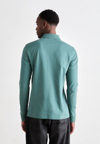 Cămașă polo cu mânecă lungă din bumbac teal, cu guler clasic și tiv drept, asociată cu blugi gri închis.