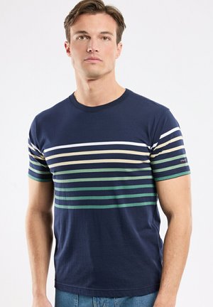 Jeune homme portant une chemise à manches courtes bleu marine avec des rayures horizontales blanches, beiges et vertes sur la poitrine et les manches, debout devant un fond uni.