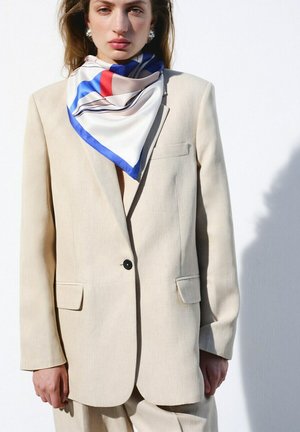 Mujer con blazer beige de gran tamaño y pantalones a juego, complementados con una bufanda blanca con rayas azules y rojas y pendientes plateados.
