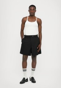 Schwarze, maßgeschneiderte Shorts mit einer vorderen Falte, Taschen und einem kontrastierenden Gürteldetail, kombiniert mit einem weißen Tanktop und gestreiften Socken.