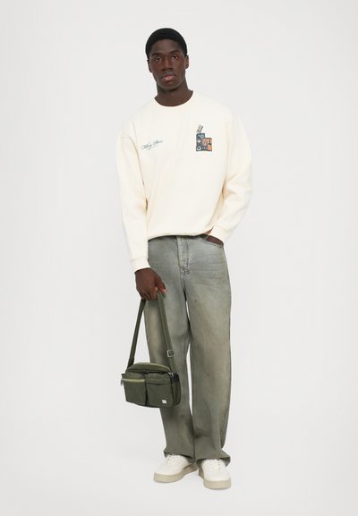Homem com um sweatshirt creme com um grafismo, jeans largos verdes desbotados, ténis brancos, segurando uma mala de ombro verde-oliva contra um fundo branco.