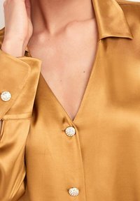 Blusa in satin dorato con scollo a V e bottoni rotondi lucidi. Presenta un colletto strutturato e una texture liscia. Cuciture dettagliate visibili sulle maniche.