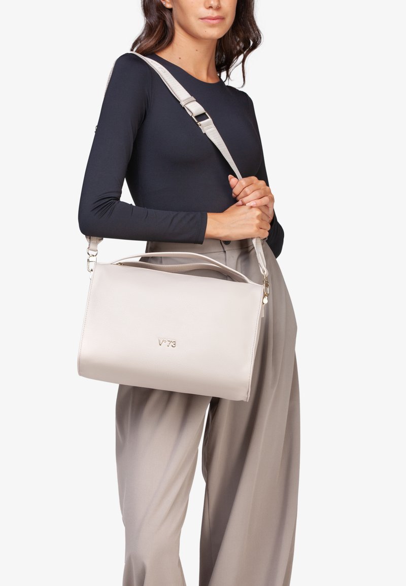 Bolso de cuero gris claro con forma estructurada, asas cortas y una correa de hombro ajustable. Presenta un sutil detalle de logotipo en la parte frontal.