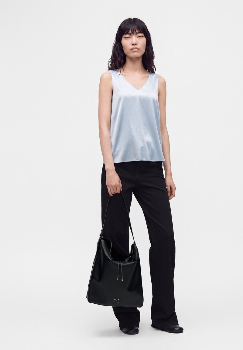 Femme debout portant un haut sans manches en satin bleu clair et un pantalon noir, tenant un grand sac à main noir dans une main.