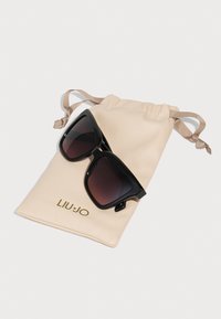 Sorte solbriller med gradientlinser, hvile på en beige stoffpose. Posene har snorer og "LIU·JO"-logo i gull.