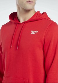 Sweat-shirt rouge avec capuche, fabriqué en tissu doux. Il présente une poche kangourou à l'avant et un logo Reebok blanc sur la poitrine gauche.