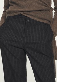 I pantaloni neri a righe mostrano una vestibilità su misura, tasche laterali e una chiusura con un solo bottone, abbinati a un maglione in maglia marrone testurizzata.
