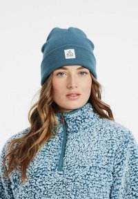 Berretto a maglia blu con orlo ripiegato e un'etichetta quadrata. Sotto, una giacca in pile azzurro chiaro con zip e finitura testurizzata.