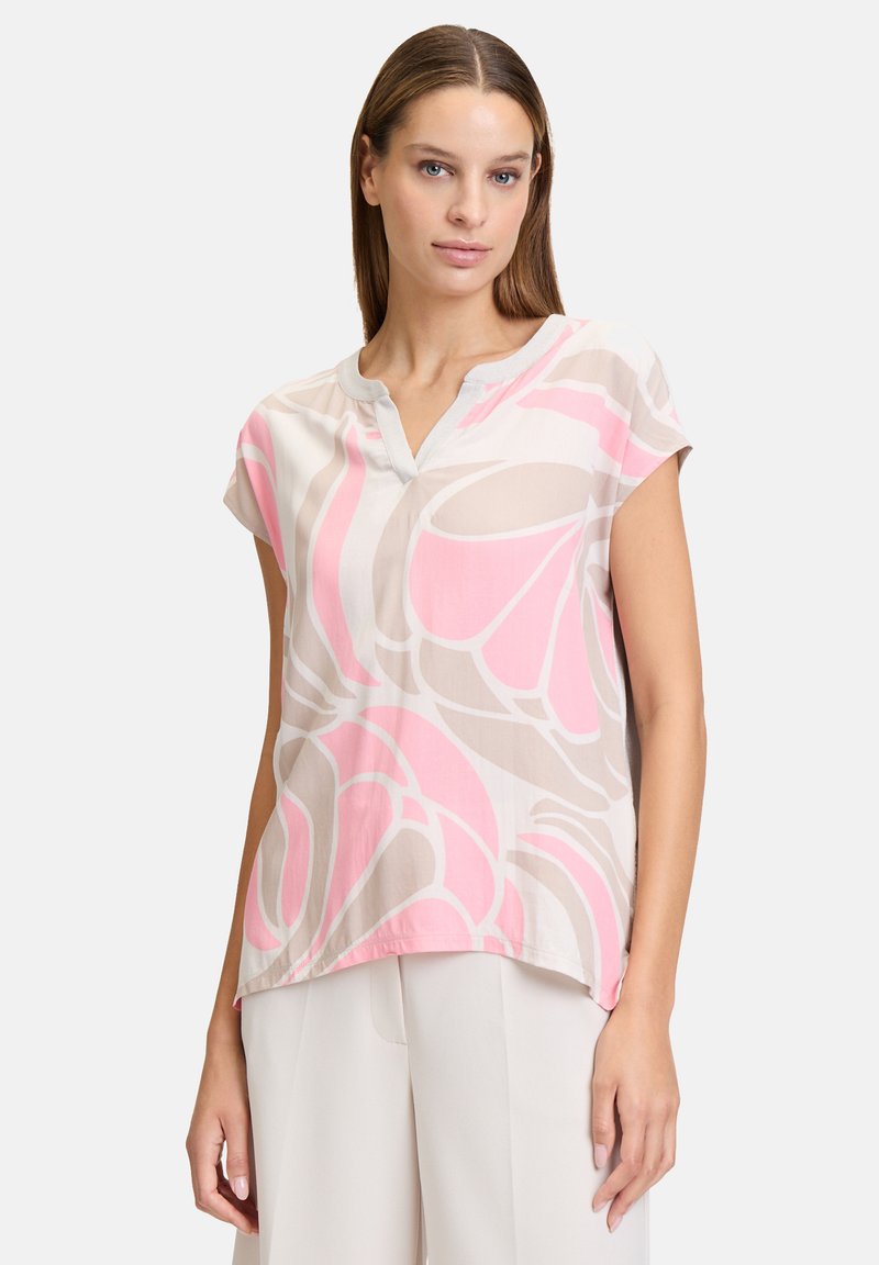 Bluzka z krótkim rękawem z lekkiego materiału, z różowym kwiatowym wzorem na beżowym tle, z dekoltem w kształcie litery V i zaokrąglonym dołem.