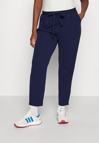 Navy blue tapered pants with a front tie, featuring side pockets and a smooth texture, paired with white sneakers and blue accents.
