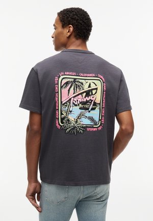 Man wearing dark gray T-shirt with colorful tropical graphic of palm trees, sailboat, and text "Tommy" and locations on the back.
