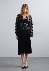 Musta paljettitoppi läpikuultavasta kankaasta, jossa pitkät hihat, sekä laskostettu musta hame, jossa on halkiokaareva helma. Yhdistetty mustiin korkokenkiin.