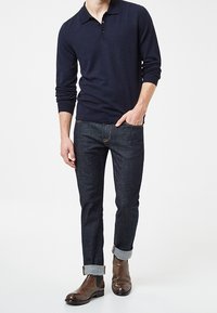 Camisa polo de manga larga en color navy, jeans de mezclilla oscuros ajustados, con los tobillos remangados, combinados con botas de cuero marrón hasta el tobillo.