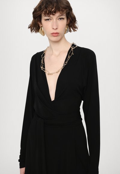 Svart omlottklänning med långa ärmar, djup V-ringning och sidoband. Halslinningen pryds av ett guldkedjehalsband. Slät tyg; minimalistisk design.