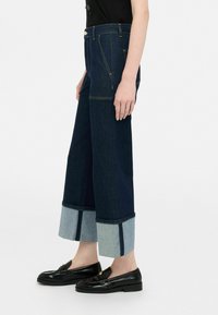 Jeans de denim de pierna ancha en azul oscuro con un puño azul claro, que presentan costuras en contraste y bolsillos delanteros. Combinados con mocasines negros.
