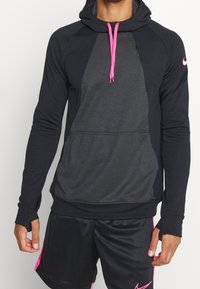 Mężczyzna w czarnej i ciemnoszarej bluzie z kapturem, z różowymi sznurkami, oraz czarnych krótkich spodenkach sportowych z różowym wykończeniem i logo.