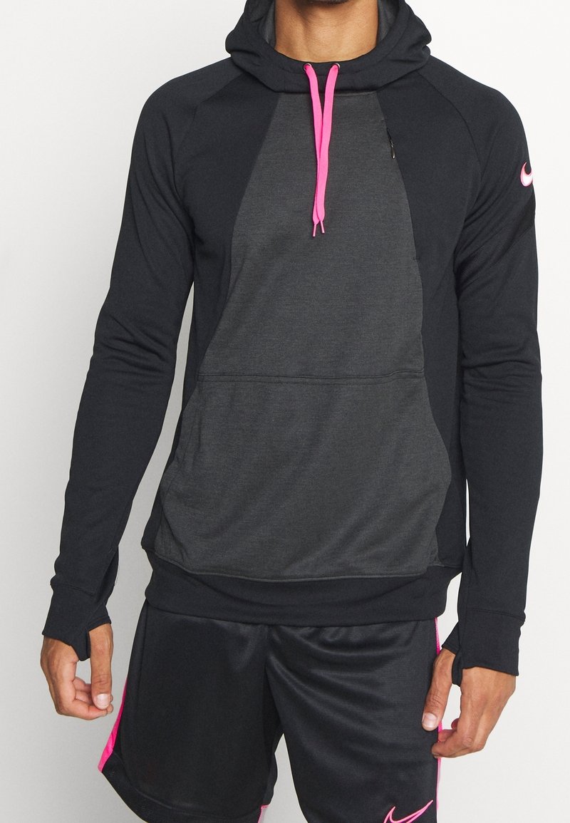 Mężczyzna w czarnej i ciemnoszarej bluzie z kapturem, z różowymi sznurkami, oraz czarnych krótkich spodenkach sportowych z różowym wykończeniem i logo.