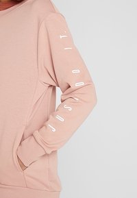 Sweatshirt rose avec des manches longues, présentant un texte blanc sur le bras, fabriqué dans un tissu doux et texturé. Poche kangourou à l'avant.