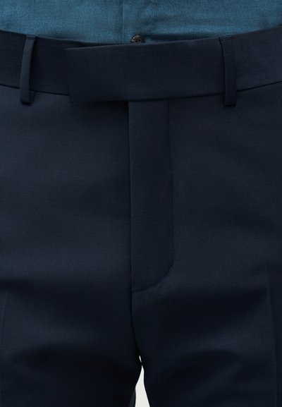 Κοντινό πλάνο στη μέση ενός σκούρου μπλε στενού παντελονιού με θηλιές για τη ζώνη και κρυφό κούμπωμα με γάντζο και μπάρα πάνω από ένα μπλε πουκάμισο.