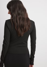 Schwarzes Langarmoberteil aus dehnbarem Stoff. Enganliegendes Design mit abgerundetem Saum und glatter Textur, kombiniert mit enganliegenden schwarzen Hosen.