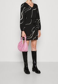 Femme portant une robe noire à lignes blanches abstraites, des bottes noires montantes jusqu'aux genoux, tenant un petit sac à main rose froncé à ses côtés.
