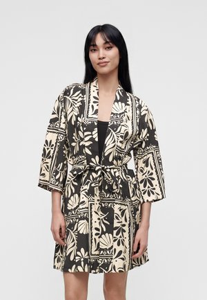 Kvinde iført sort og beige kimono-stil badekåbe med blomsterprint, bundet om taljen med trekvartærmer mod en ensfarvet baggrund.