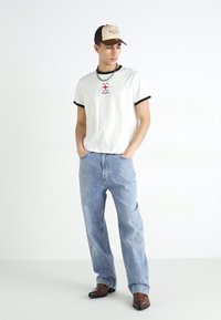 Valkoinen T-paita mustilla reunoilla, jossa on punainen logo; vaaleansiniset suorat farkut; tummanruskeat cowboy-kengät; beige lippalakki logolla.